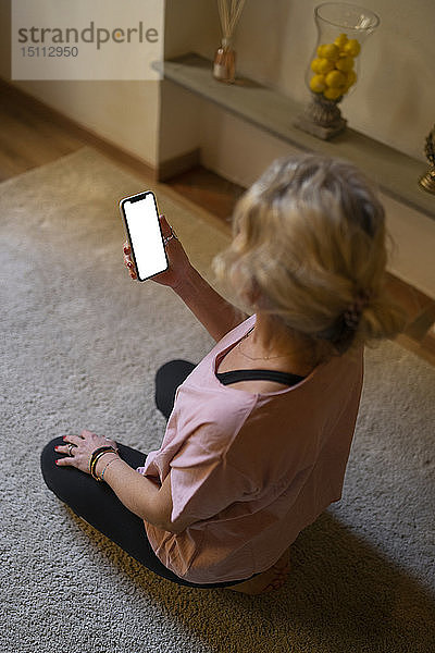 Reife Frau sitzt auf einem Teppich im Wohnzimmer und schaut auf das Display ihres Mobiltelefons