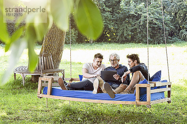 Männer einer Familie sitzen auf einem Schaukelbett im Garten  unterhalten sich  benutzen ein digitales Tablet