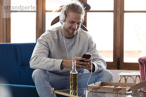 Lächelnder junger Mann sitzt mit Handy und Kopfhörern auf der Couch