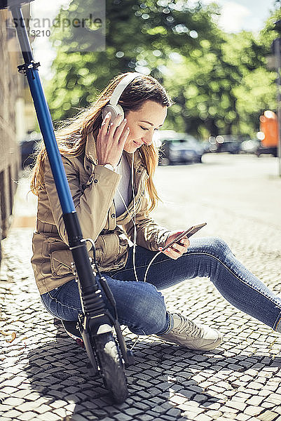 Lächelnde Frau sitzt auf einem E-Scooter und hört Musik mit Kopfhörern und Smartphone