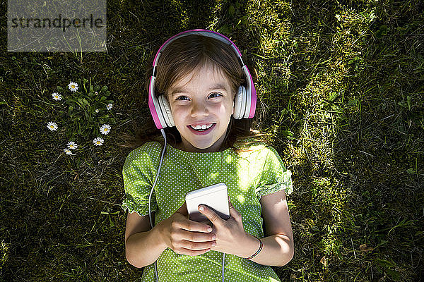 Porträt eines kleinen Mädchens  das auf einer Wiese liegt und mit Kopfhörer und Smartphone Musik hört