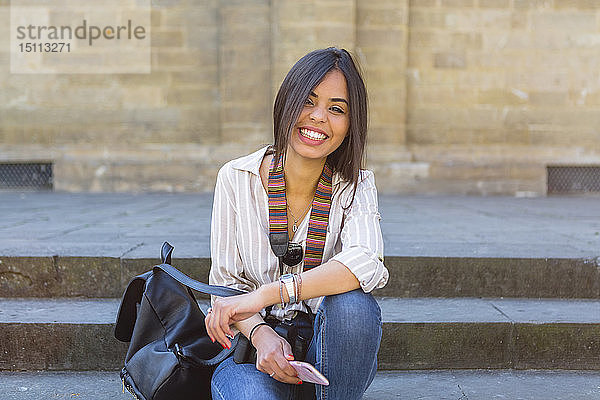 Italien  Florenz  Porträt eines glücklichen jungen Touristen  der mit Rucksack und Smartphone auf einer Treppe sitzt