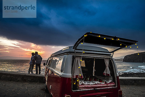 Älteres Ehepaar reist in einem Oldtimer-Van und küsst sich bei Sonnenuntergang am Meer