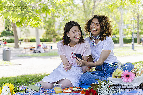 Zwei lachende Frauen mit Handy und Kopfhörern im Park