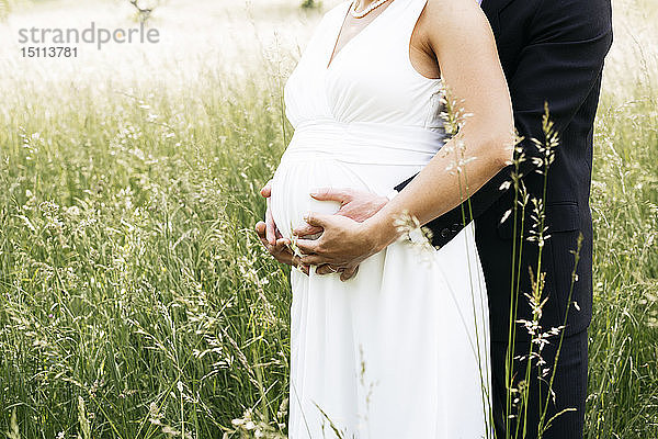 Schwangere Braut mit ihrem Ehemann hält Babybauch auf einer Wiese