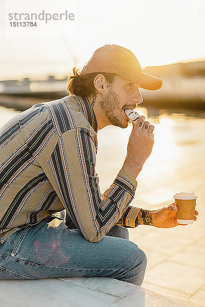 Tätowierter Mann mit Kaffee zum Donut-Essen bei Sonnenuntergang
