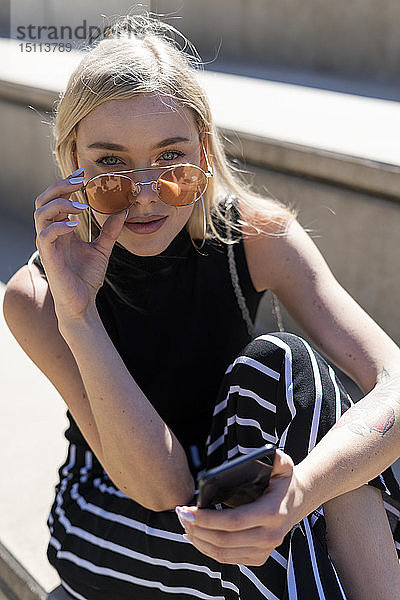 Porträt einer modischen jungen Frau  die auf einer Treppe im Freien sitzt und eine Sonnenbrille trägt