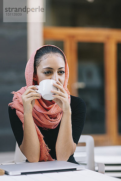 Junge Frau mit Laptop und Kopftuch in einem Straßencafé