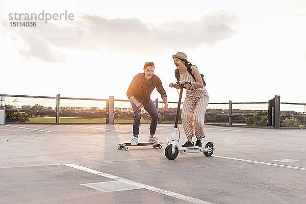 Junger Mann und Frau fahren auf Longboard und Elektroroller auf dem Parkdeck bei Sonnenuntergang