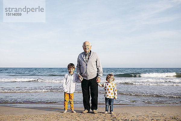 Porträt eines glücklichen Grossvaters  der mit seinen Enkelkindern Hand in Hand am Strand steht
