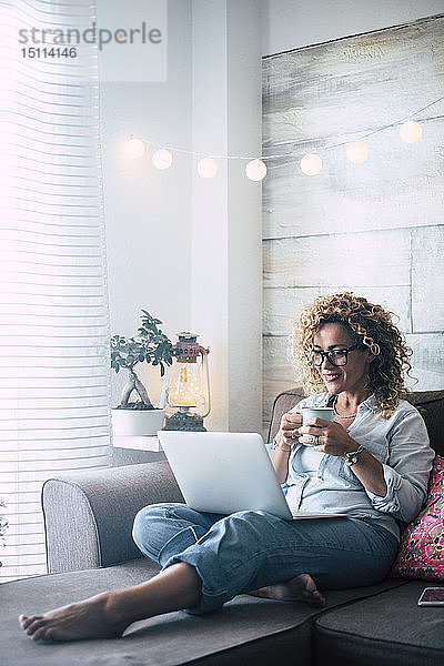 Frau auf Couch zu Hause mit Kaffeetasse und Laptop