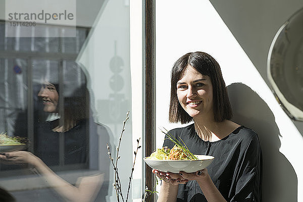 Lächelnde junge Frau steht bei Sonnenschein am Fenster und hält einen Teller Salat