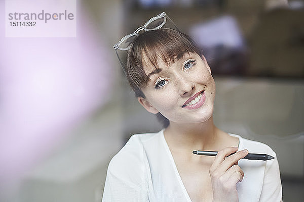 Porträt einer lächelnden jungen Geschäftsfrau mit Brille und Bleistift hinter Glasscheibe