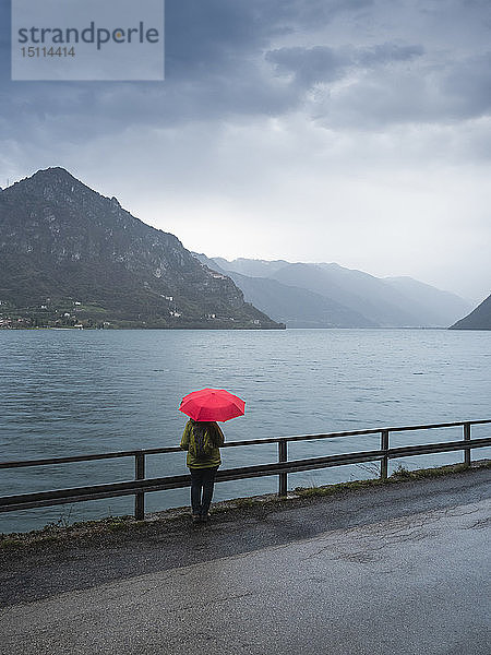 Italien  Lombardei  Rückenansicht einer älteren Frau mit Rucksack und rotem Regenschirm mit Blick auf den Idrosee