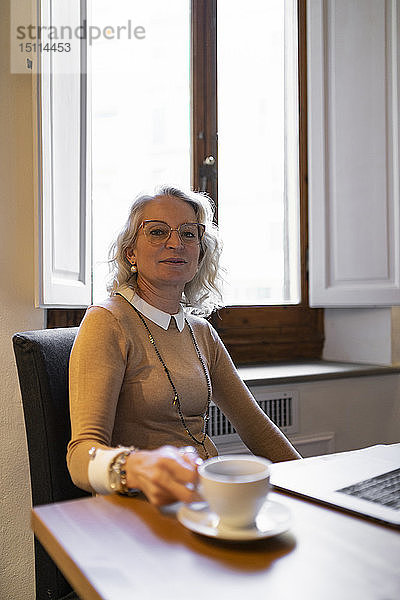 Porträt einer zufriedenen reifen Geschäftsfrau  die mit Laptop und einer Tasse Kaffee am Tisch sitzt