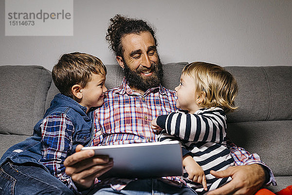 Glücklicher Vater sitzt mit seinen Kindern auf der Couch und benutzt digitales Tablet
