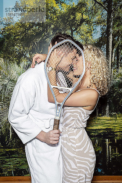 Junges Paar steht vor einer Fototapete  küsst sich hinter einem Tennisschläger
