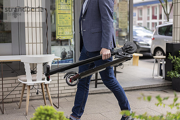 Geschäftsmann mit gefaltetem E-Scooter beim Gehen auf dem Bürgersteig  Teilansicht