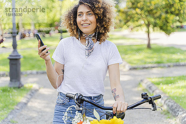 Lächelnde Frau mit Handy  Kopfhörern und Fahrrad im Park