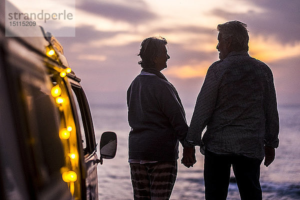 Älteres Ehepaar reist in einem Oldtimer-Van und betrachtet den Sonnenuntergang am Meer