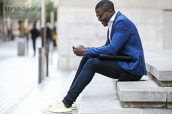 Junger Geschäftsmann in blauer Anzugjacke  auf Schritt und Tritt sitzend und mit Smartphone