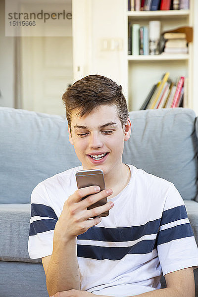 Porträt eines lächelnden Teenagers  der mit seinem Handy vor der Couch sitzt