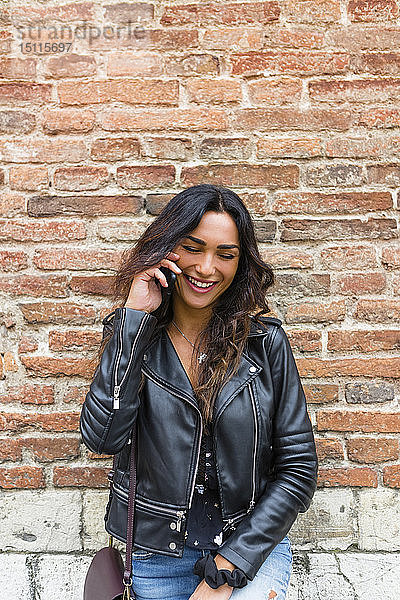 Porträt einer jungen Frau in schwarzer Lederjacke  mit Smartphone  im Hintergrund Backsteinmauer