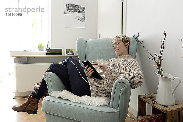 Entspannte Frau sitzt zu Hause im Sessel und benutzt Tablette