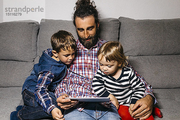 Vater sitzt mit seinen Kindern auf der Couch und sieht sich Filme auf einem digitalen Tablet an