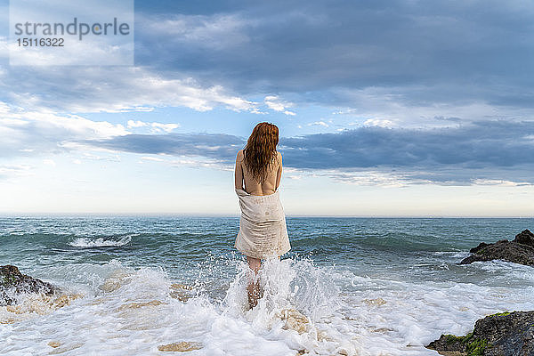 Rückenansicht einer rothaarigen  hemdlosen jungen Frau  die vor dem Meer steht