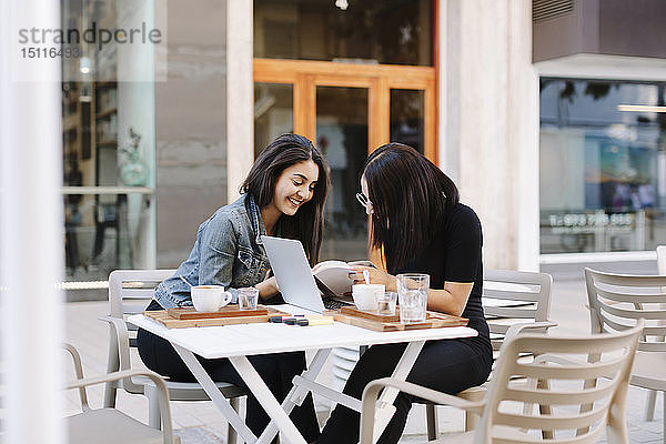 Zwei Freunde sitzen zusammen mit Buch und Laptop in einem Straßencafé