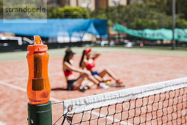 Orangefarbene Trinkflasche mit Tennisspielerinnen  die im Hintergrund auf dem Platz sitzen