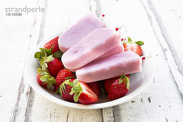 Hausgemachte Erdbeer-Joghurt-Eislollys mit frischen Erdbeeren in einer Schale auf weißem Holz