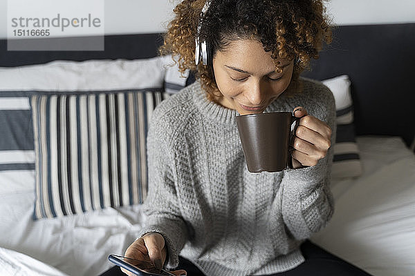 Frau sitzt auf dem Bett  trinkt Kaffee  hört Musik mit Kopfhörern und Smartphone