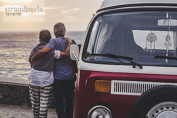 Ein älteres Ehepaar reist in einem Oldtimer-Van und schaut mit den Armen auf das Meer