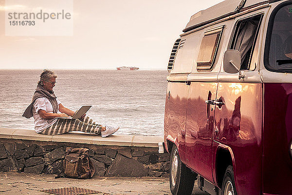 Ältere Frau  die mit einem Oldtimer-Van reist  auf einer Mauer am Meer sitzt und einen Laptop benutzt
