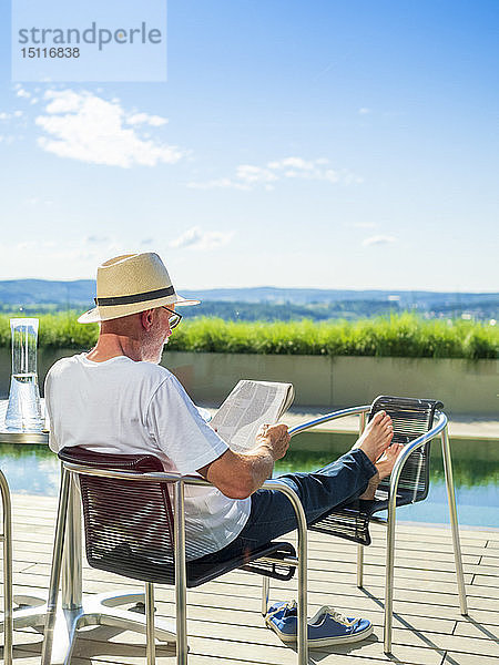 Rückansicht eines älteren Mannes  der Zeitungspapier liest und am Schwimmbad sitzt