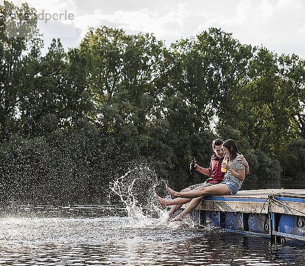 Junges Paar trinkt und planscht mit Wasser auf einem Steg an einem abgelegenen See