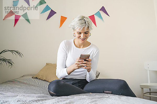 Lächelnde Frau  die zu Hause am Bett ein Mobiltelefon benutzt