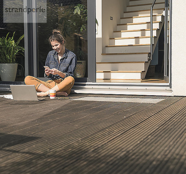 Glückliche junge Frau sitzt zu Hause auf der Terrasse und benutzt Handy und Laptop