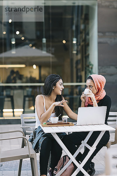 Zwei Freunde sitzen mit Laptop in einem Straßencafé zusammen und unterhalten sich