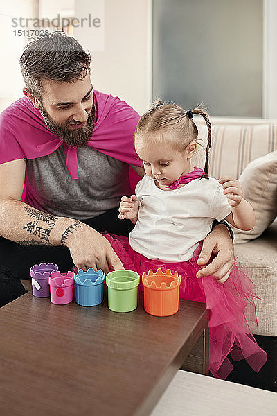 Vater und Tochter im Superhelden- und Superfrau-Kostüm  spielen mit gestapelten Tassen