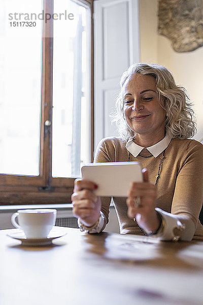 Porträt einer lächelnden reifen Geschäftsfrau  die mit einer Tasse Kaffee am Tisch sitzt und auf ihr Handy schaut