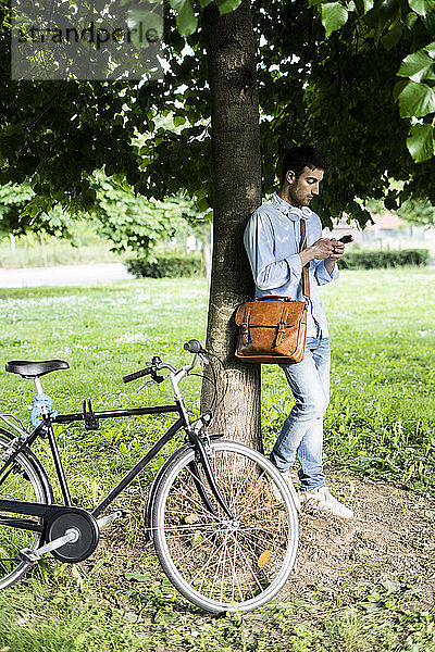 Junger Mann mit Fahrrad und Smartphone  Kopfhörer um den Hals  unter einem Baum stehend