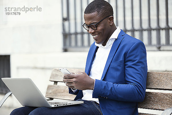 Junger Geschäftsmann in blauer Anzugjacke sitzt auf Bank und benutzt Smartphone