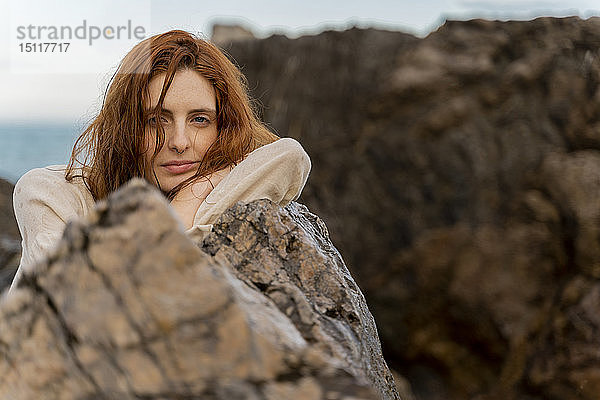 Porträt einer rothaarigen jungen Frau mit Nasenpiercing  die sich an einen Felsen lehnt