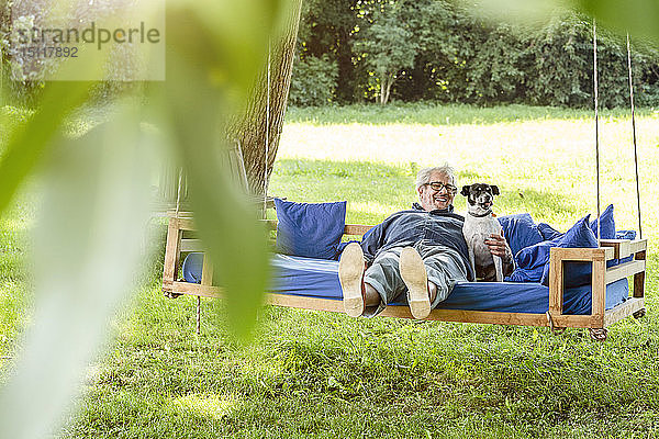 Älterer Mann entspannt sich auf einem Schaukelbett in seinem Garten mit seinem Hund