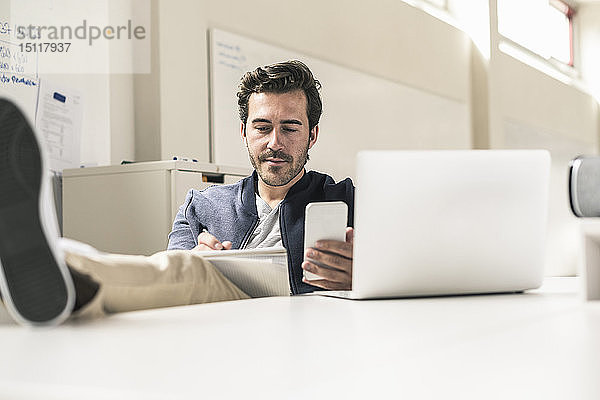 Junger Geschäftsmann  der entspannt in einem modernen Büro mit Smartphone und Laptop arbeitet