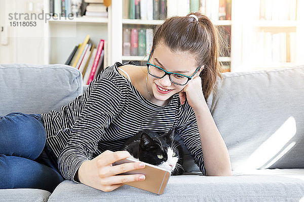 Lächelndes Mädchen  das zu Hause auf der Couch liegt und mit der Katze Selfie nimmt
