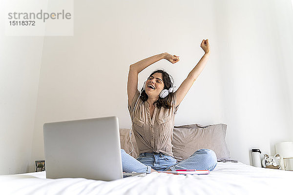 Glückliche junge Frau sitzt mit Kopfhörern und Laptop auf dem Bett